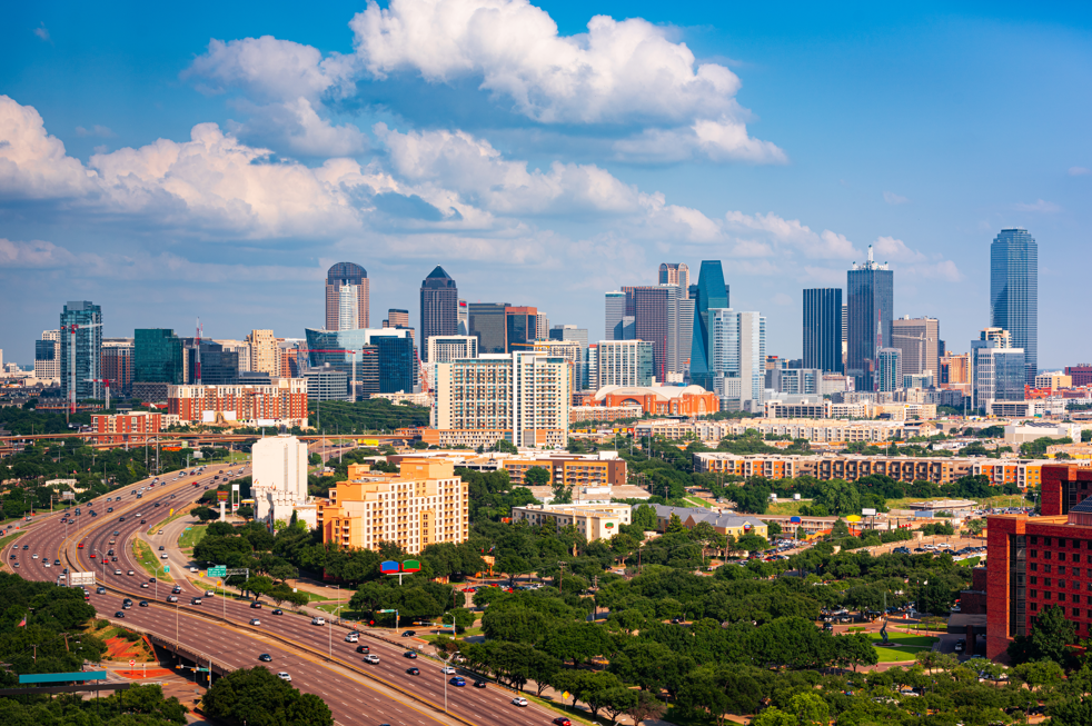 Downtown Dallas skyline with high-rise towers, highway traffic, and green trees under blue sky